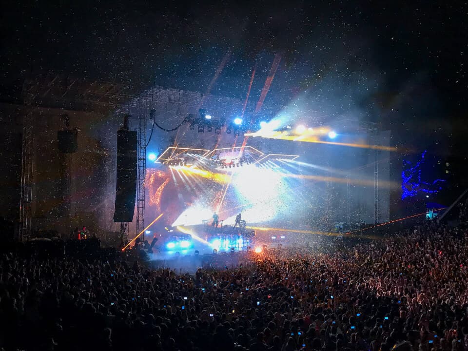 Rig lighting dan line array tergantung pada truss panggung terbuka, sorot cahaya menembus malam di atas penonton